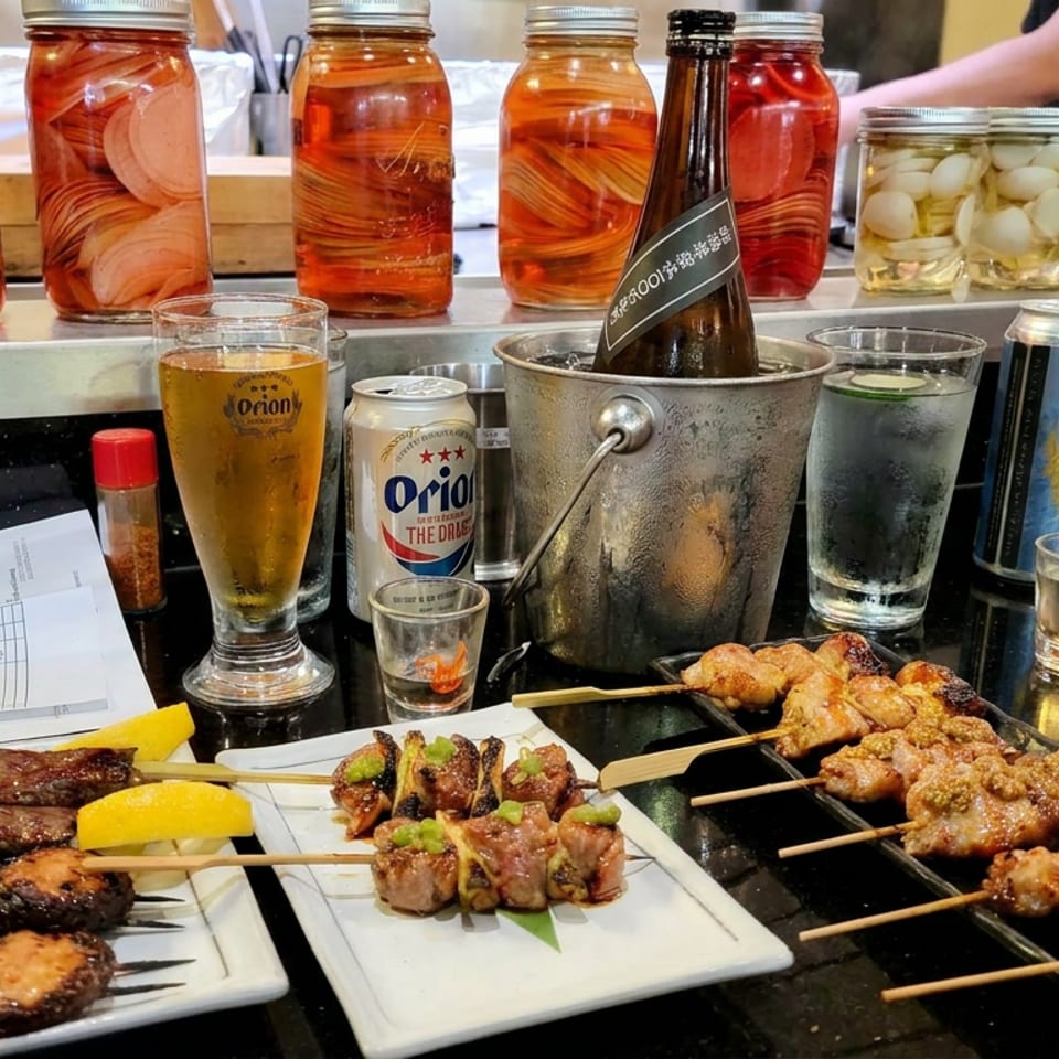 Assorted grilled skewers, beer, and pickled vegetables fill a restaurant counter beside a chilled bottle and dipping dishes.
