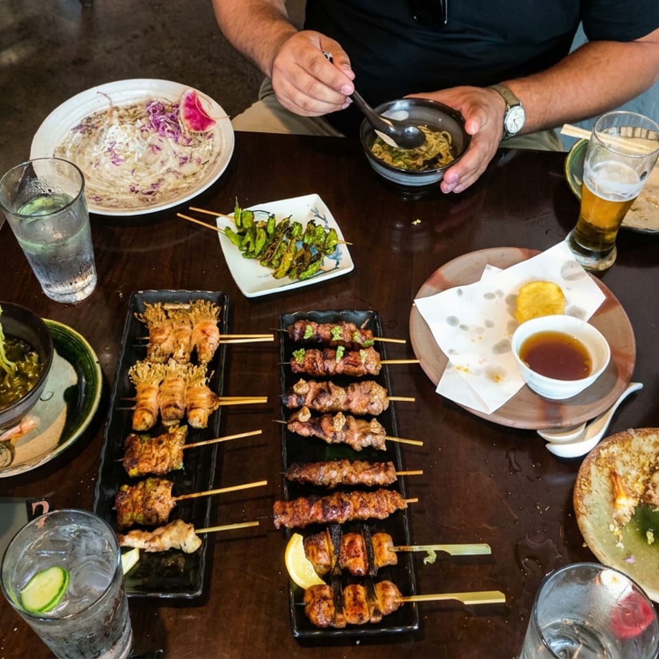 Diners share Japanese noodles, grilled skewers, dumplings, and drinks around a dark restaurant table.