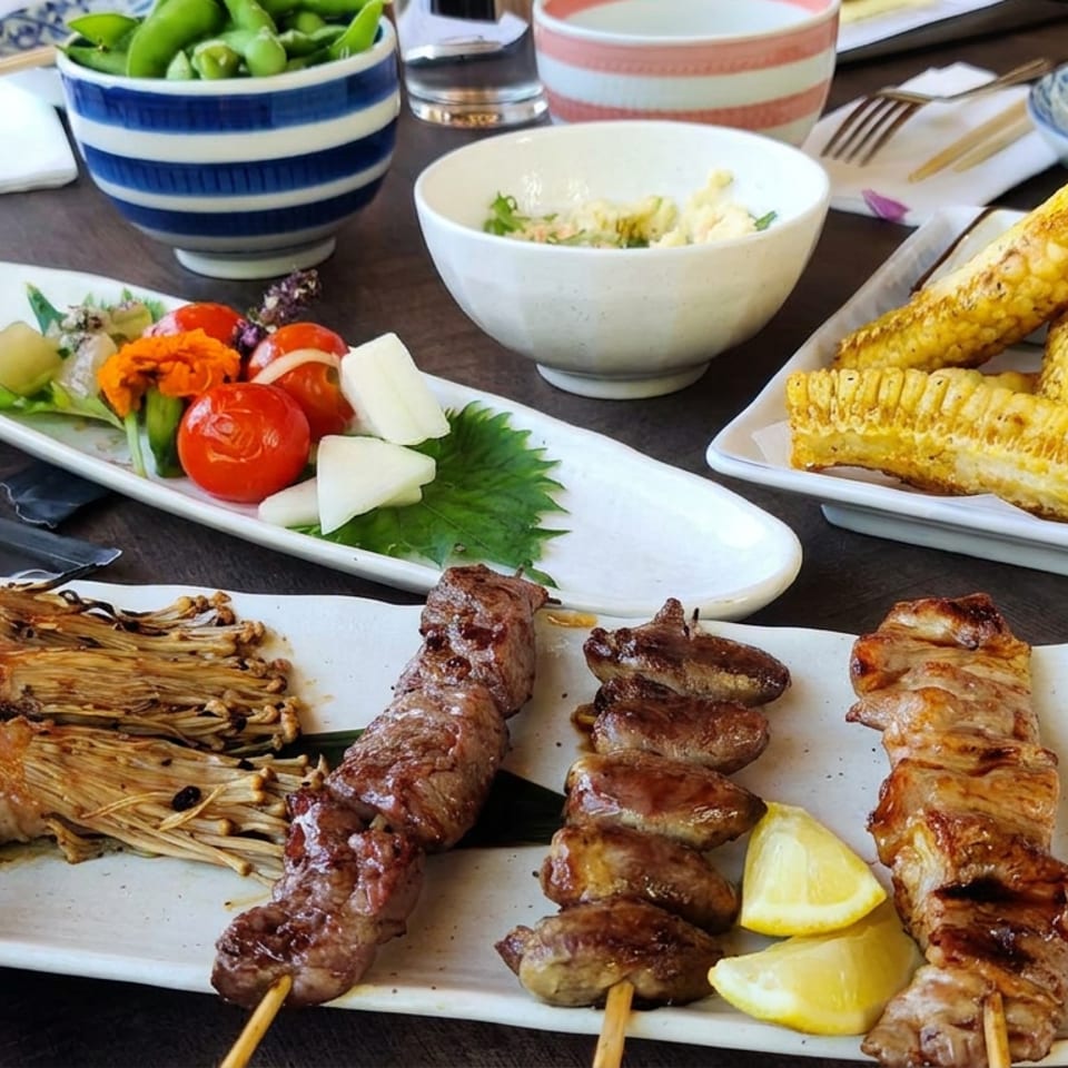 Japanese-style meal with grilled skewers, bacon-wrapped mushrooms, vegetables, fried corn, rice, and patterned dishes on a restaurant table.