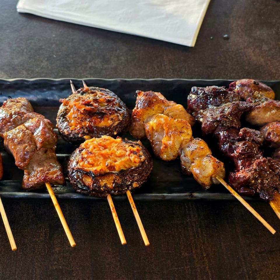 Black platter featuring grilled sausages, meat skewers, stuffed mushrooms, roasted vegetables, and lemon wedges.