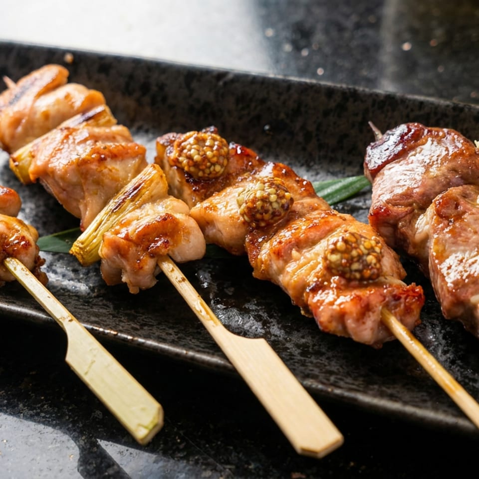 Glazed grilled meat skewers with mustard seeds, leeks, and a lemon wedge arranged on a black rectangular plate.