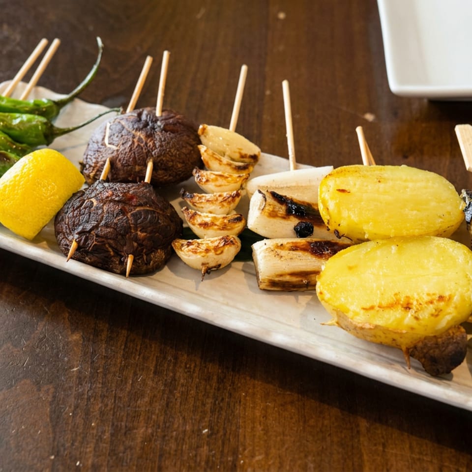 Grilled vegetable skewers with peppers, mushrooms, onions, eggplant, potatoes, and lemon arranged on a white platter.