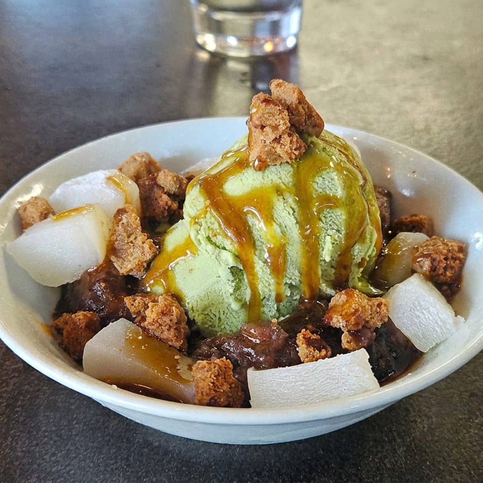 Bowl of green pandan ice cream topped with caramel, crispy crumbs, dark jelly, and chewy white cubes.