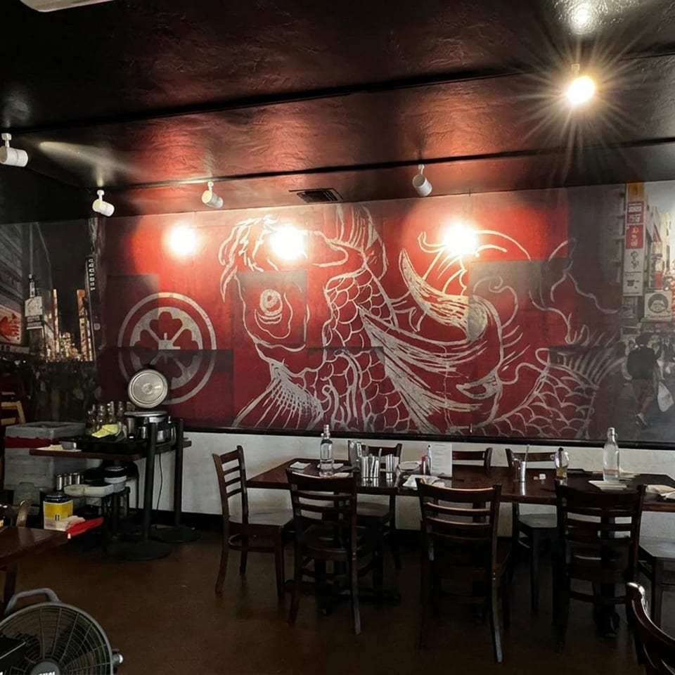 Japanese restaurant interior with dark wooden tables, a red dragon mural, beverage refrigerator, and street-facing windows.