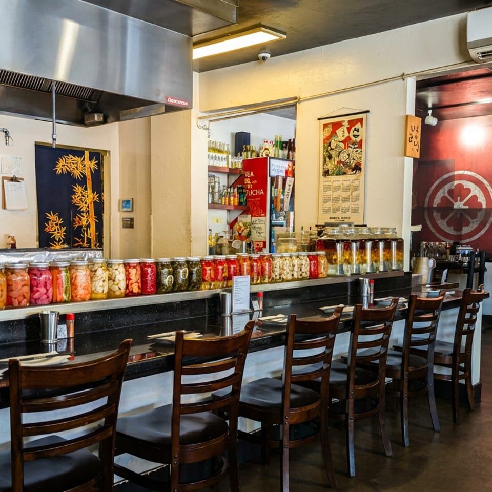 Japanese restaurant interior with colorful pickled vegetables in jars, counter seating, open kitchen, and a large red fish mural.